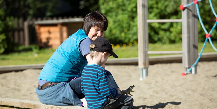 En vuxen ler mot ett barn. De sitter på kanten av en sandlåda.