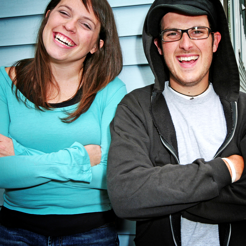 En man med glasögon och en grå tröja står bredvid en kvinna i blå tröja. Det båda står med armarna i kors framför en ljusblå vägg och skrattar tillsammans. 