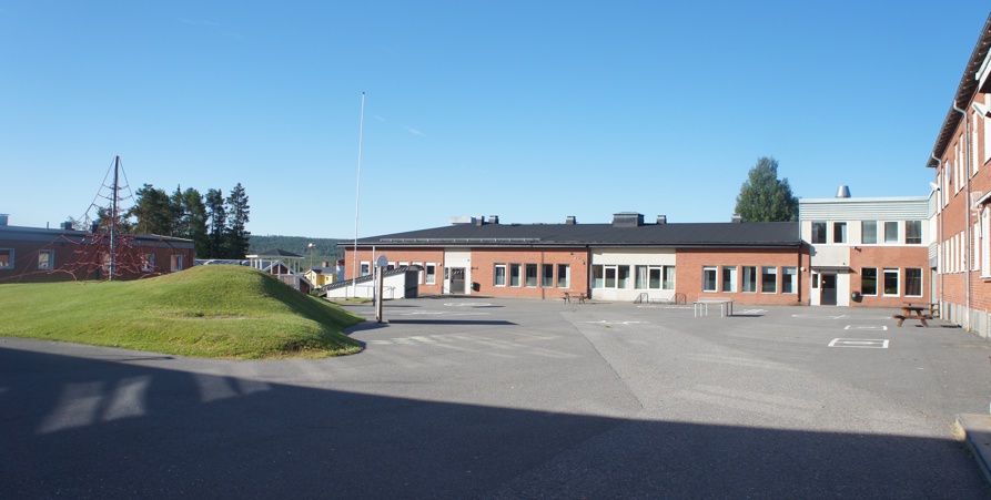 Sommarbild tagen in mot mellanstadiets skolgård med asfalterad gårdsplan, gräskulle och byggnader.