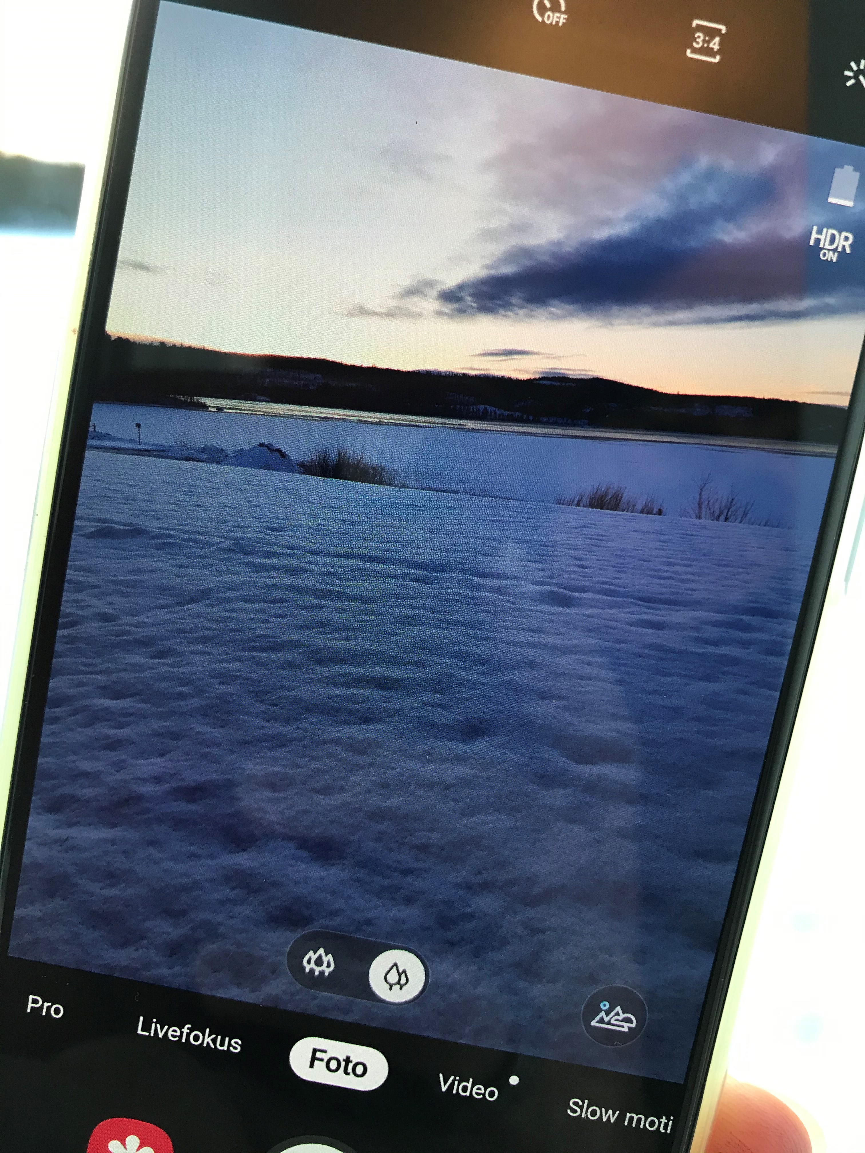 Närbild på mobiletelefon som tar ett foto på en sjö med snö och is. Berg i bakgrunden.